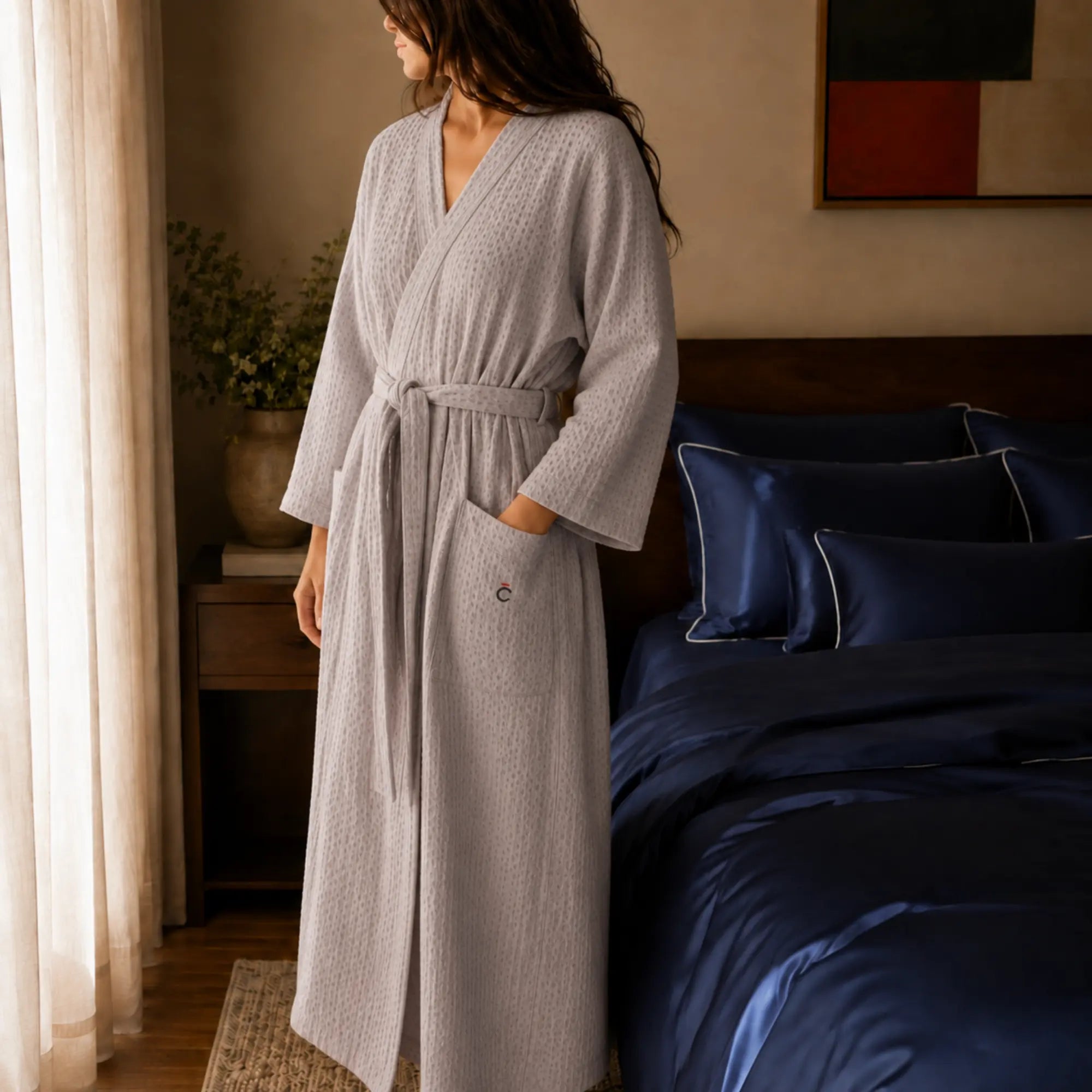 Femme portant le peignoir Waffle en coton gaufré gris perle Maison CÉTOILE dans une chambre au linge de lit bleu nuit.