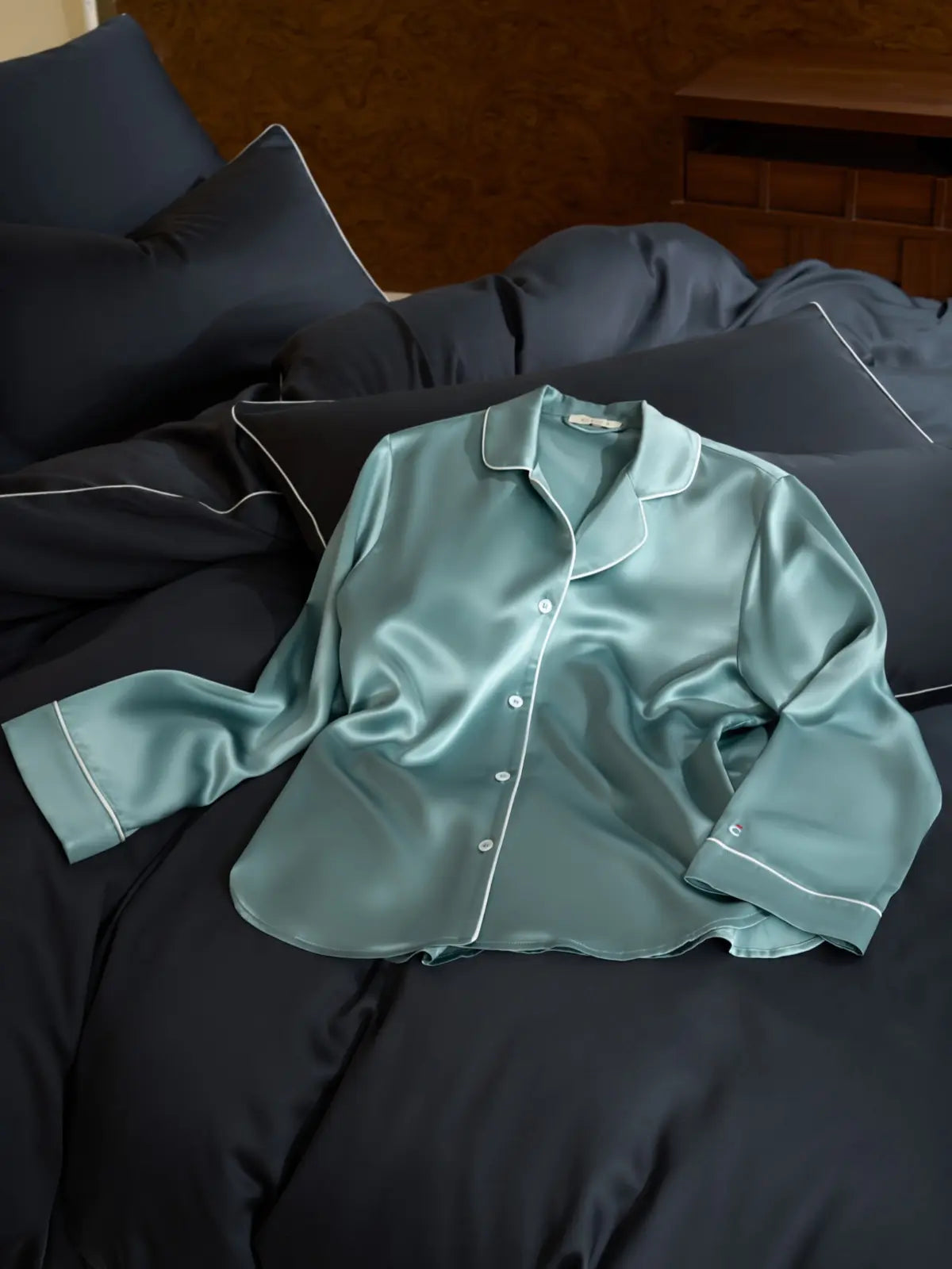 Haut de pyjama en soie de mûrier bleu grisé Brume d’Été, avec passpoil blanc, posé sur un lit bleu nuit.